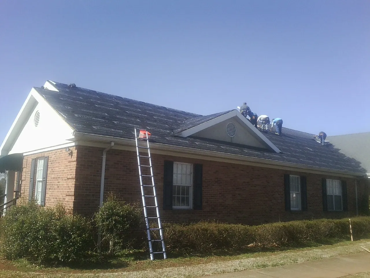 Crew tearing shingles off a brick ranch roof with a ladder set — Emergency Roof Repair work by our crew in Pendergrass