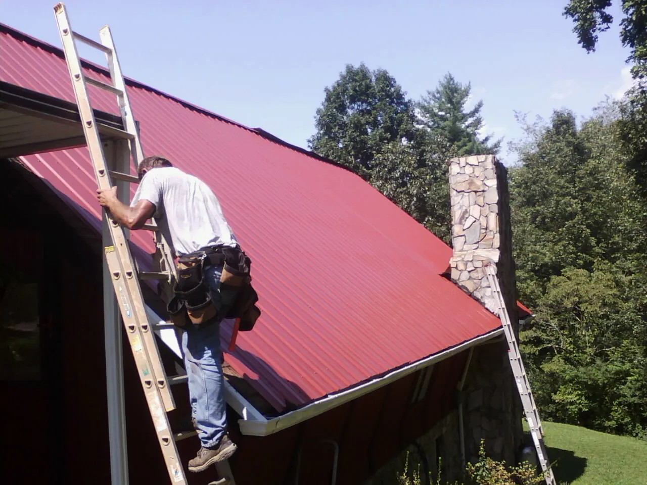 Roofer on a ladder beside a red metal roof and stone chimney — Soffit & Fascia Repair by Roofing Brothers in Pendergrass